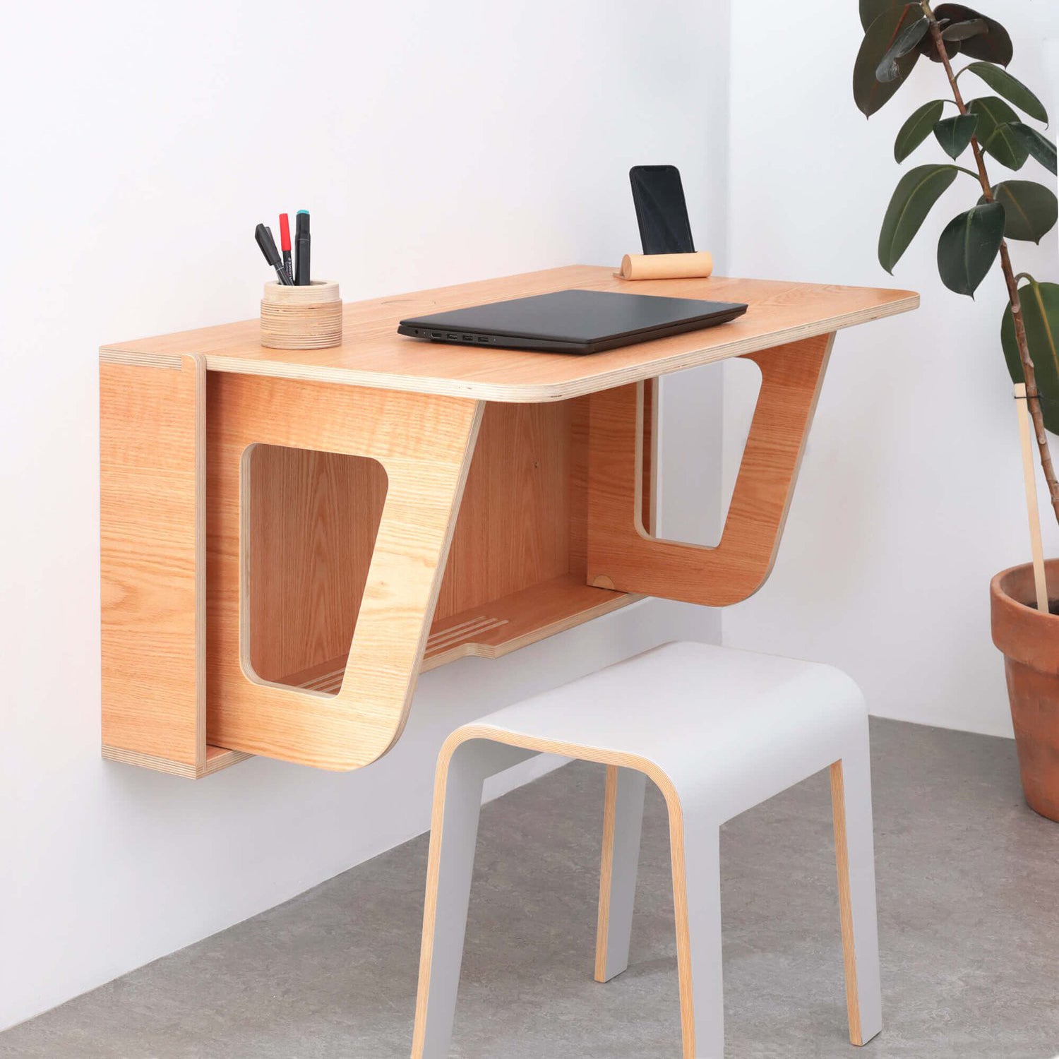 A modern wall-mounted Leaf Desk made of wood with a laptop and stationery on it, alongside a white chair and decorative plants in the background.